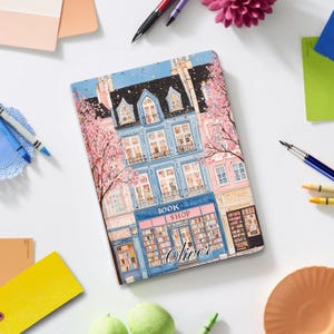 May include: A notebook featuring a detailed illustration of a book shop, with the name "Oliver" displayed. The building is painted in blue and pink hues, with cherry blossom trees. The words "BOOK SHOP" are above the shop's entrance.