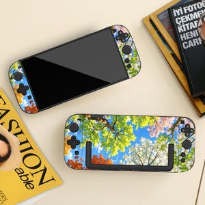 Puede incluir: Dos consolas de juegos portátiles con una piel vibrante con diseño de dosel forestal. La piel muestra hojas de otoño verdes, blancas y naranjas contra un cielo azul brillante. Las consolas tienen pantallas, botones y joysticks negros.