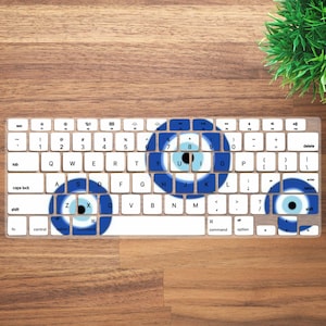 Op de afbeelding: Een wit toetsenbord met blauwe en witte evil eye-ontwerpen op de toetsen. Het toetsenbord ligt op een houten oppervlak met een kleine groene plant in de rechterbovenhoek. Het toetsenbord heeft standaard toetsen.