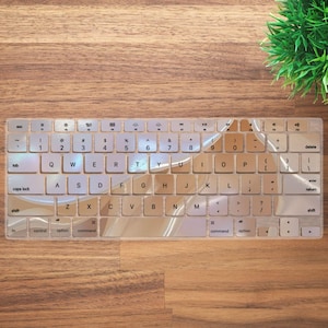 Puede incluir: Una cubierta de teclado transparente con un diseño de mármol en tonos marrones y blancos. La cubierta está diseñada para adaptarse a un teclado Apple estándar y presenta todas las teclas necesarias, incluidas las teclas de función, las teclas de flecha y las teclas numéricas.