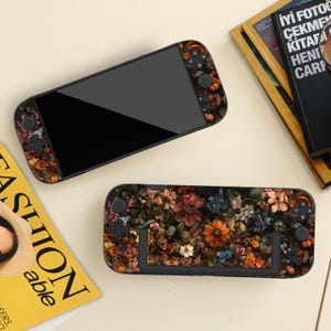 Könnte beinhalten: Zwei schwarze Handheld-Spielkonsolen mit floralen Designs. Eine Konsole ist geöffnet und zeigt den Bildschirm, während die andere geschlossen ist. Das Blumenmuster zeigt eine Mischung aus orangefarbenen, braunen und blauen Blumen.