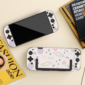 May include: Two white and black gaming console skins with a space-themed design. One skin is on a console, the other is separate. The design includes rockets, stars, and planets. The consoles have black screens and buttons.
