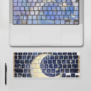 以下が含まれることがあります： 青と金の三日月と星の模様が描かれた天体テーマのキーボードカバーが付いたシルバーのノートパソコン。その下に、お揃いの外付けキーボードスキンと黒いスタイラスペンが、明るいグレーの表面に配置されています。