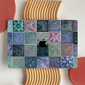 Puede incluir: Una funda decorativa para portátil con un diseño de mosaico de baldosas cuadradas en tonos azules, verdes y morados. Cada baldosa muestra una imagen en relieve única, incluyendo una mariposa, una estrella de mar y motivos florales. El logotipo de Apple es visible.