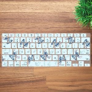 May include: A keyboard cover featuring a light blue and white design with the character Stitch from Lilo & Stitch. The keys are white with black lettering and Stitch graphics. The cover is on a wooden surface.