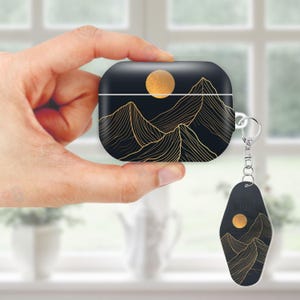 May include: Black AirPod case with a gold mountain and sun design. A matching keychain with the word "MOTEL" and a similar mountain and sun graphic hangs from the case. The case is held in a hand.