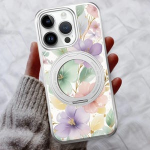 May include: Clear phone case with a floral design featuring purple, pink, and yellow flowers, green leaves, and gold accents. The case has a silver ring holder and a clear border. The phone case is held in a person's hand.