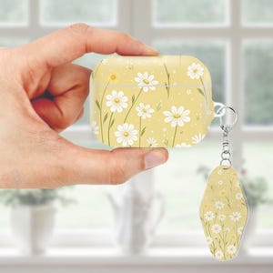 May include: Yellow AirPod case and matching keychain featuring a daisy floral pattern. The case is rectangular with rounded edges and a silver keychain attachment. The daisy design includes white flowers, green stems, and a yellow background.