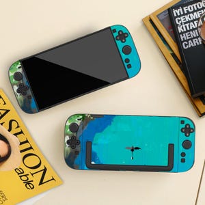 May include: Two Nintendo Switch consoles with custom skins. One console features a camouflage pattern in shades of green, blue, and brown. The other has a teal and blue watercolor design with a black dragon silhouette. Both have teal controllers.