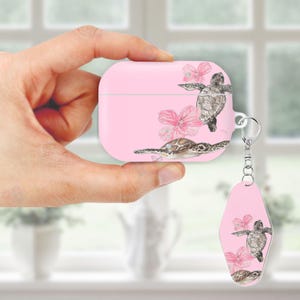 May include: Pink AirPod case and matching keychain featuring watercolor sea turtles and pink flowers. The case has a silver metal ring for the keychain. The design is repeated on the keychain.