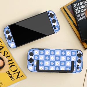 May include: Two blue and white floral patterned gaming console cases. One case is attached to a black screen, and the other is separate. The cases have a checkerboard pattern with white flowers. A magazine is also visible.