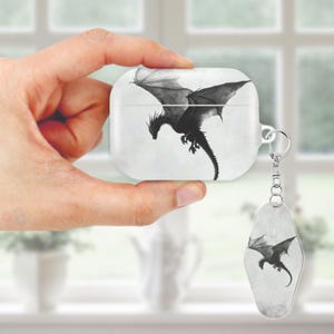 May include: White AirPods case and matching keychain with a black dragon illustration. The dragon is shown with outstretched wings against a cloudy backdrop. The case has a silver metal loop for attaching the keychain.