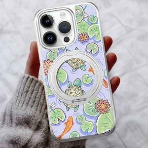 May include: Clear phone case with a whimsical pond design. The case features turtles, koi fish, lily pads, and dragonflies on a light purple background. A silver ring is attached to the back. The case is designed for a smartphone.