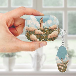 May include: A white and blue AirPods case with a pastoral scene featuring a figure in a robe, sheep, and a field of flowers. A matching keychain with the same design hangs from the case. The case is held in a hand.