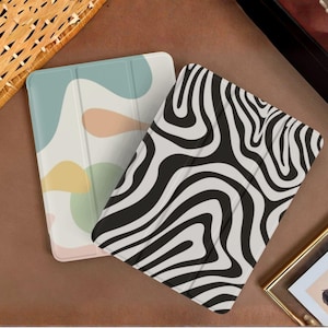 May include: Two rectangular tablet cases. One case features a soft-toned abstract pattern with muted blue, peach, and yellow organic shapes on a cream background. The other case displays a bold, high-contrast black and white wavy zebra-like pattern.