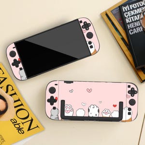May include: Two pink game console controllers with a cat paw design. The controllers have black buttons and a black screen. The design includes cat paws, hearts, and cat faces. The controllers are on a light beige surface.