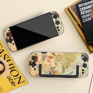 May include: Two video game consoles with a floral and fauna design. The design includes a parrot, cherries, a rabbit, and other elements on a beige background. The consoles have black screens and buttons.