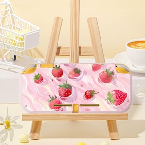 May include: A pink and white Nintendo Switch case featuring a pattern of strawberries. The case has yellow accents on the sides and is displayed on a small wooden easel. The strawberries are red with green leaves.