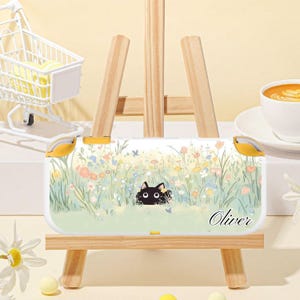 May include: A white handheld gaming console case featuring a whimsical floral design with a black cat peeking out. The case has yellow accents and the name "Oliver" is written on the bottom right.