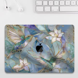 May include: A laptop skin with a stained-glass effect, depicting hummingbirds and flowers in blue, green, and gold. The design includes an Apple logo. A white keyboard is visible in the background.
