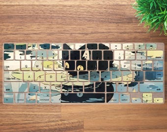 May include: A keyboard cover with a cat face design in shades of blue, green, and black. The cat's eyes are yellow. The keys are visible through the design. The cover is designed to fit a standard keyboard layout.