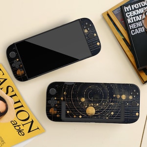 Peut inclure: Deux consoles de jeux portables noires avec un design céleste. L'une est face vers le haut, affichant un écran noir, tandis que l'autre est face vers le bas, présentant un motif de système solaire doré et noir. Les consoles sont sur une surface claire.