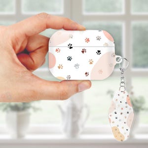 May include: White AirPod case and matching keychain featuring a repeating pattern of paw prints in various colors, including black, brown, and pink. The case has a rounded rectangular shape and a silver keychain attachment.