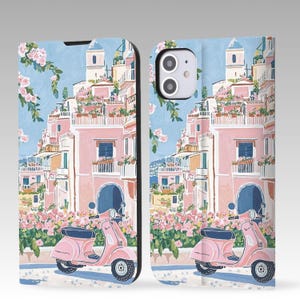 May include: A phone case with a colourful illustration of an Italian coastal town. The design features pink buildings, flowering plants, and a pink scooter. The background is a light blue sky, creating a scenic view.
