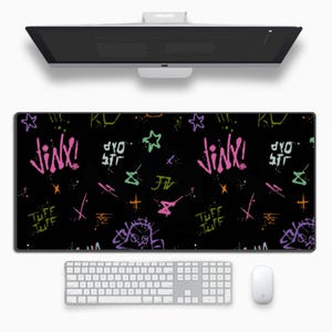 Puede incluir: Alfombrilla de escritorio rectangular negra con diseños coloridos de estilo grafiti, incluyendo la palabra "Jinx!" en rosa y blanco. La alfombrilla presenta estrellas, formas y el texto "Tuff Tuff". Un teclado y un ratón blancos están debajo.