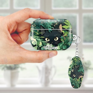 May include: A green and black AirPod case and keychain featuring a black cat peeking through foliage. The name "Oliver" is written in cursive on the case. The design has a whimsical, nature-inspired theme.