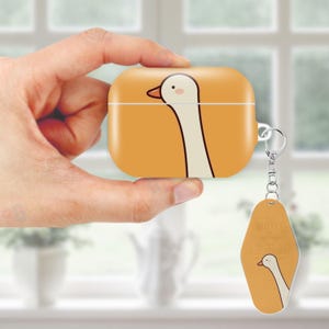 May include: A yellow AirPod case featuring a cartoon goose design. The case is held in a hand and has a matching keychain with the words "MOTEL EL PASO" and a goose illustration. The background is a blurred interior scene.