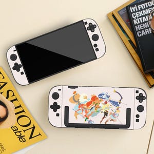 May include: Two white and black Nintendo Switch consoles. One has a blank black screen, the other a colourful graphic design. The consoles are on a light surface, with a magazine and book in the background.