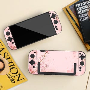 May include: Two pink Nintendo Switch consoles with a floral design. The consoles have a black screen and black buttons. The floral design includes pink cherry blossoms. The consoles are on a light beige surface.
