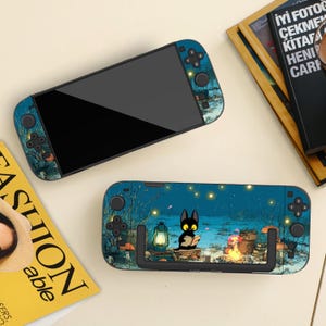 May include: Two handheld gaming consoles with a blue and green forest design. One console is face up, the other is face down, displaying a black cat reading a book by a campfire. The consoles have black screens and control buttons.