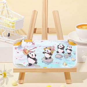 May include: A white Nintendo Switch case featuring a whimsical panda design. The case has a light blue background with cartoon pandas, flowers, and water elements. The case is displayed on a small wooden easel.