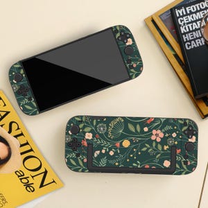 Peut inclure: Deux consoles de jeux portables avec un motif floral vert foncé. Les consoles ont un écran noir et des boutons de commande. Le motif floral comprend des fleurs roses, jaunes et blanches avec des feuilles vertes. Les consoles sont sur une surface beige clair.