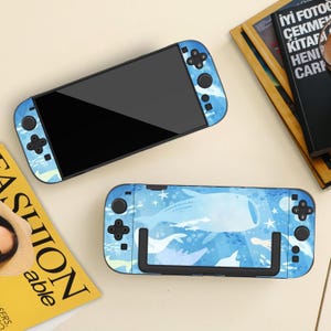 以下が含まれることがあります： クジラと海のテーマのデザインが施された、青と白の2つのNintendo Switchコンソール。コンソールは、クジラ、魚、星のパターンで装飾されています。コントローラーには黒いボタンが付いています。雑誌と本が背景にあります。