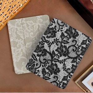Puede incluir: Dos fundas rectangulares para lector electrónico con intrincados patrones de encaje. Una funda muestra un diseño de encaje blanco sobre un fondo beige claro, mientras que la otra presenta un patrón de encaje negro sobre fondo blanco.