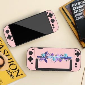 Peut inclure: Deux consoles Nintendo Switch roses. L'une est unie, l'autre présente un motif floral et une épée. Les consoles ont des écrans et des boutons noirs. L'arrière-plan comprend un magazine et un livre.