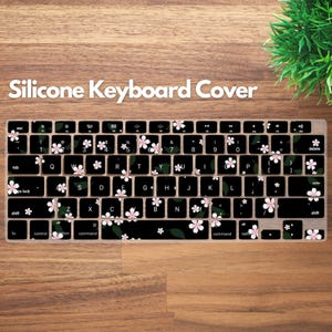 May include: A black silicone keyboard cover with a floral design. The cover features white and pink cherry blossom flowers and green leaves. The text "Silicone Keyboard Cover" is at the top.