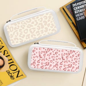 Op de afbeelding: Twee witte draagtassen met een handvat, met een luipaardprint. De ene tas heeft een beige en crèmekleurig patroon, terwijl de andere een roze en mauve patroon heeft. De koffers zijn rechthoekig en lijken voor elektronica te zijn.