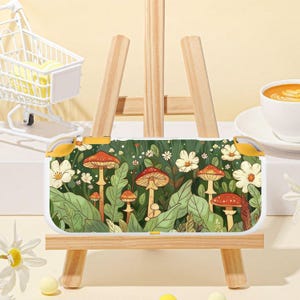 May include: A white and yellow handheld gaming console case featuring a whimsical mushroom and floral design. The case has a white border and yellow buttons. The background is a lush green with red-capped mushrooms and white flowers.