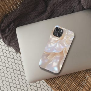 May include: A phone case with a pearlescent, abstract design in white and cream hues. The case is placed on a silver laptop, with a dark grey blanket and a woven basket in the background. The case has a glossy finish and a swirling pattern.