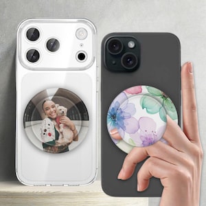May include: Two phone cases with attached pop sockets. One case is clear with a photo of a person holding a dog. The other is dark gray with a floral design in shades of blue, pink, and green.