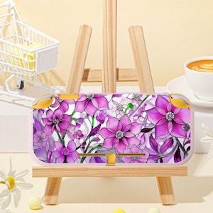 May include: A pink and purple floral patterned Nintendo Switch Lite case. The case features a design of stylized flowers in shades of pink and purple, with white accents. The case is displayed on a small wooden easel.