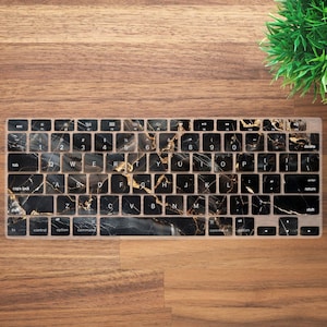 Può includere: Una cover per tastiera con motivo marmorizzato nero e oro. La cover ha uno sfondo nero con venature dorate, che copre i tasti. La cover per tastiera è su una superficie di legno.