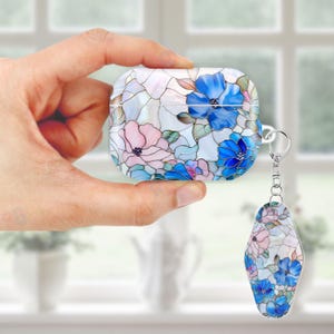 May include: White AirPod case and matching keychain with a stained glass floral design. The case features blue and pink flowers with green leaves, set against a white background. The keychain mirrors the case's design.