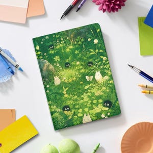 May include: A rectangular notebook featuring a vibrant green forest floor illustration with small white creatures, black soot sprites, and a butterfly. The cover art depicts lush foliage, clover leaves, and tiny white flowers in a dense, grassy setting.