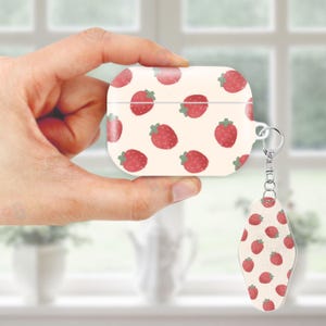 May include: A white AirPod case and matching keychain featuring a repeating pattern of red strawberries with green leaves. The case has a silver metal ring for attaching the keychain. The background is a soft cream color.