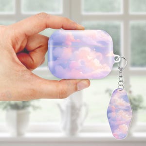 Puede incluir: Una funda lila para AirPods con un diseño de nubes en tonos rosa, morado y blanco. Tiene un llavero plateado y una etiqueta a juego con el tema de las nubes. La funda está sujeta en una mano.